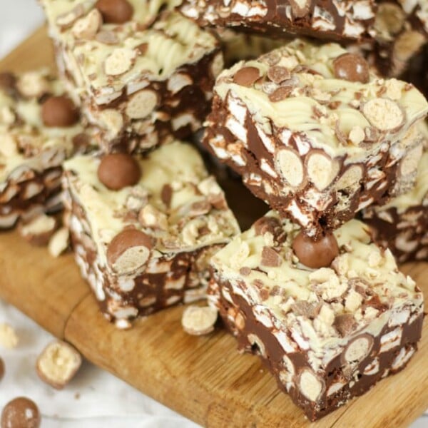 Side angle photo of a pile of Malteser Rocky Road pieces on a wooden board with pack of Maltesers in the background.