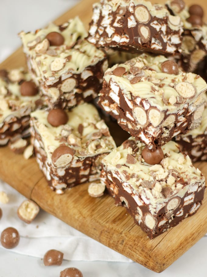 Side angle photo of a pile of Malteser Rocky Road pieces on a wooden board with pack of Maltesers in the background.