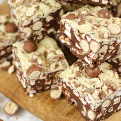 Side angle photo of a pile of Malteser Rocky Road pieces on a wooden board with pack of Maltesers in the background.