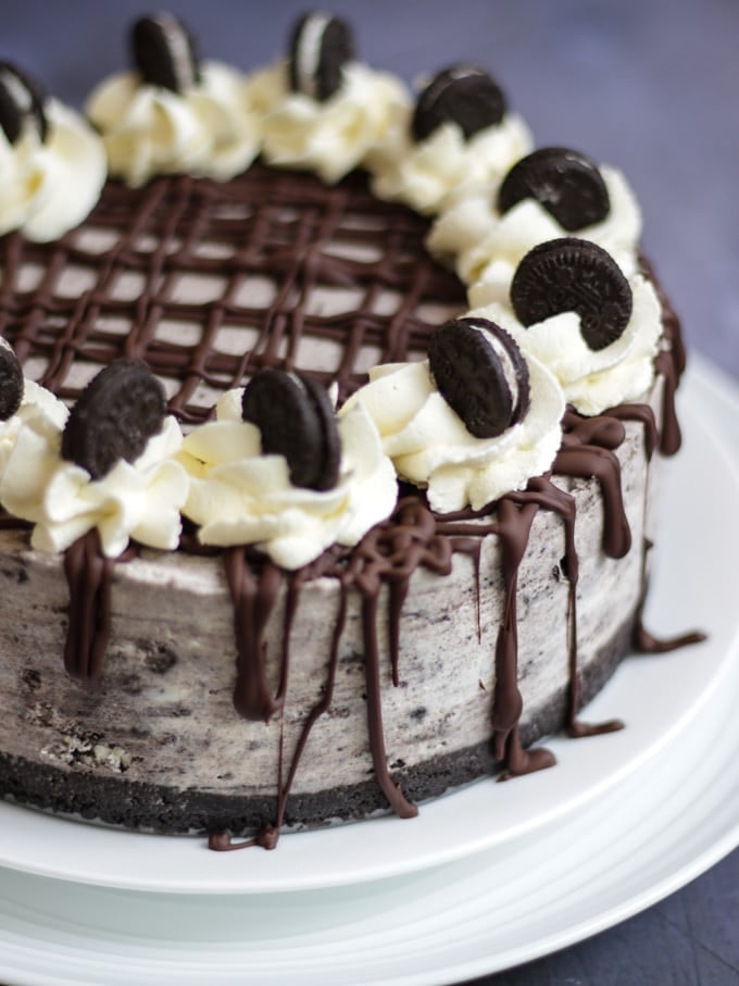 Side angle photo of a no bake Oreo cheesecake on two white plates. Drizzled with dark chocolate, topped with piped whipped cream and topped with mini Oreos and a pack of Oreo cookies in the background on dark grey background.