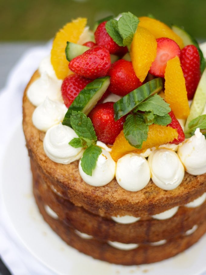 Overhead angled photo of Pimms cake topped with piped buttercream icing, cucumber, strawberries, orange segments and mint on a white plate in a garden with green grass in background to demonstrate pimms cake recipe.