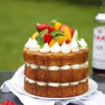 Side photo of Pimms cake topped with piped buttercream icing, cucumber, strawberries, orange segments and mint on a white plate in a garden with green grass in background to demonstrate pimms cake recipe.