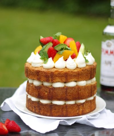 Side photo of Pimms cake topped with piped buttercream icing, cucumber, strawberries, orange segments and mint on a white plate in a garden with green grass in background to demonstrate pimms cake recipe.