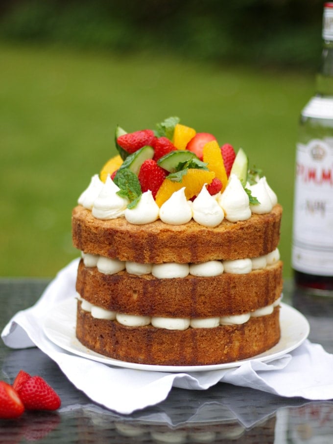 Side photo of Pimms cake topped with piped buttercream icing, cucumber, strawberries, orange segments and mint on a white plate in a garden with green grass in background to demonstrate pimms cake recipe.