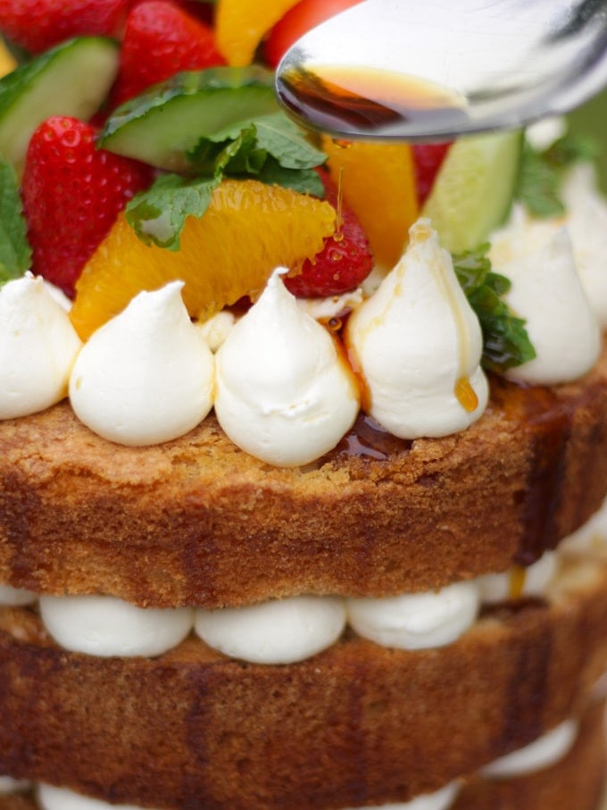 Side photo of Pimms cake with spoon drizzling pimms syrup on cake topped with piped buttercream icing, cucumber, strawberries, orange segments and mint on a white plate in a garden with green grass in background to demonstrate pimms cake recipe.