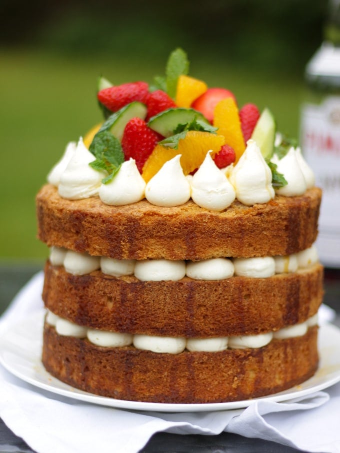 Side photo of Pimms cake topped with piped buttercream icing, cucumber, strawberries, orange segments and mint on a white plate in a garden with green grass in background to demonstrate pimms cake recipe.