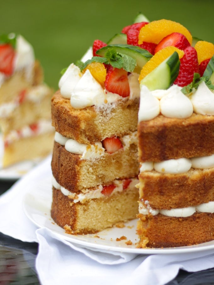 Side photo of Pimms cake topped with piped buttercream icing, cucumber, strawberries, orange segments and mint on a white plate in a garden with green grass in background to demonstrate pimms cake recipe.