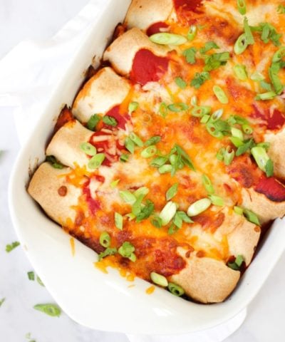 Overhead photo of chicken enchiladas in a white rectangular dish with tomato sauce, cheese melted, onions (scallions) and coriander (cilantro) for easy chicken enchiladas recipe which is also slimming world friendly.