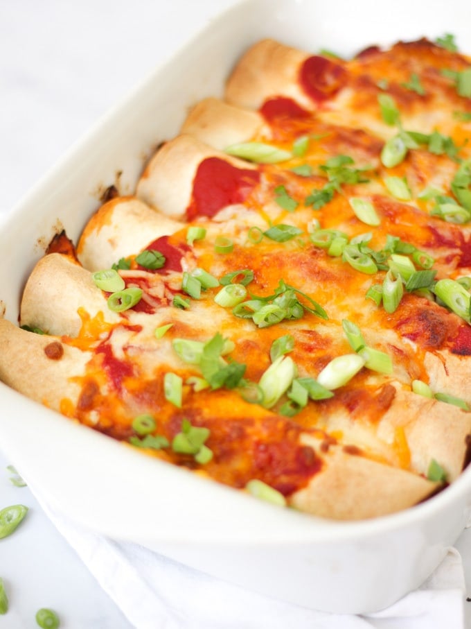 Side angle photo of chicken enchiladas in a white rectangular dish with tomato sauce, cheese melted, onions (scallions) and coriander (cilantro) for easy chicken enchiladas recipe which is also slimming world friendly.