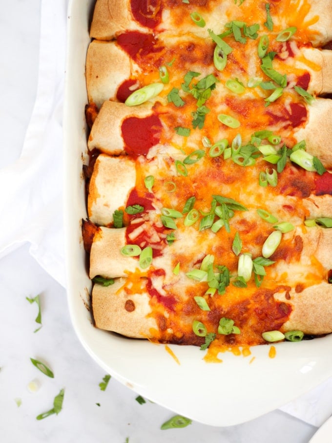 Overhead photo of chicken enchiladas in a white rectangular dish with tomato sauce, cheese melted, onions (scallions) and coriander (cilantro) for easy chicken enchiladas recipe which is also slimming world friendly.