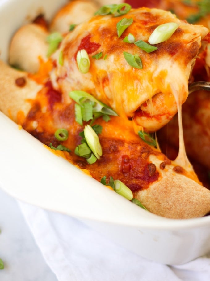 Close up view of chicken enchiladas being spooned and lifted with melted cheese in a white rectangular dish with tomato sauce, cheese melted, onions (scallions) and coriander (cilantro) for easy chicken enchiladas recipe which is also slimming world friendly.