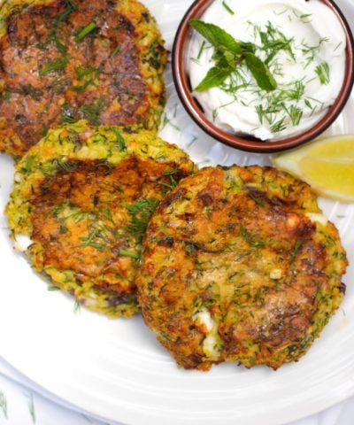 Overhead shot of courgette fritters recipe with 3 fritters on a white plate and a brown pot of yoghurt and dill dip.