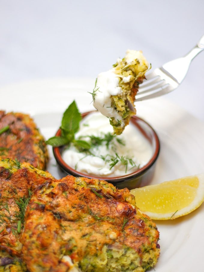 Overhead shot of courgette fritters recipe with 3 fritters on a white plate and a brown pot of yoghurt and dill dip with fork pushing in and dipping a mouthful in.