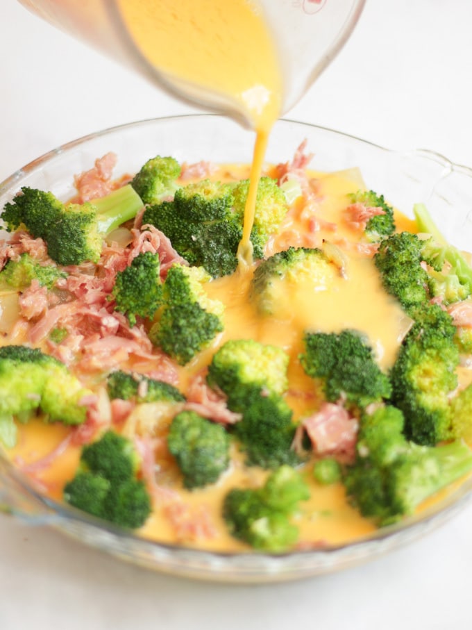 Broccoli and ham in a round glass dish for Crustless Quiche recipe with egg pouring into it.