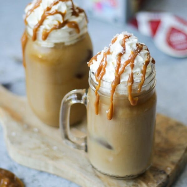 Two glass kilner mugs of iced coffee with salted caramel and tassimo pods in the background on grey background.