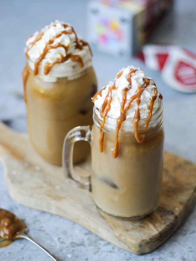 Two glass kilner mugs of iced coffee with salted caramel and tassimo pods in the background on grey background.