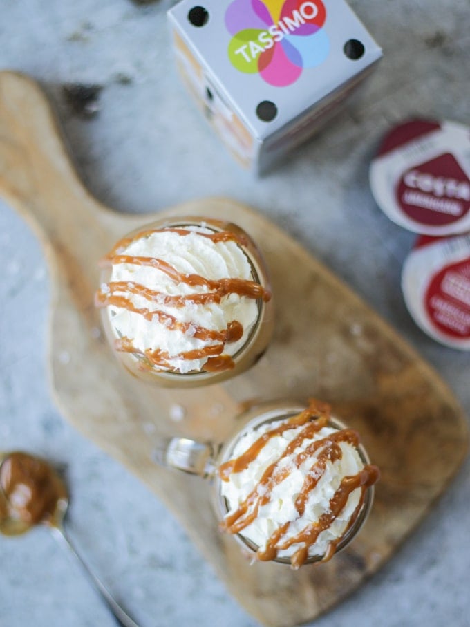 Overhead photo of two glass kilner mugs of iced coffee with salted caramel and tassimo pods in the background on grey background.