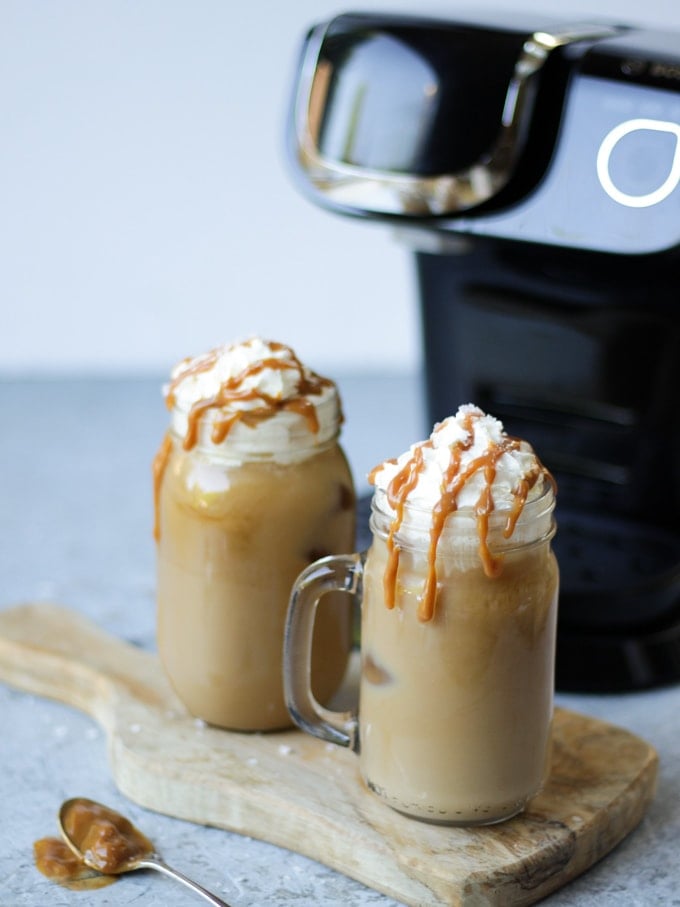 Two glass kilner mugs of iced coffee with salted caramel and tassimo pods in the background on grey background with Tassimo coffee machine in the background.