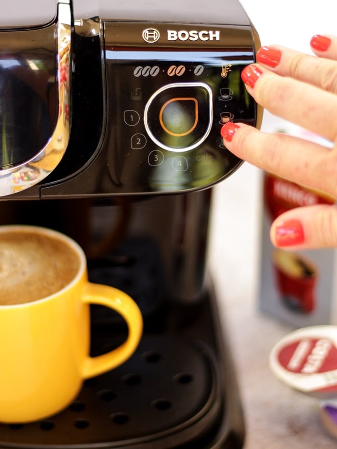 Tassimo coffee machine with finger with red fingernail pressing button with yellow mug filled with coffee and coffee pods in the background for making Iced coffee recipe with salted caramel.