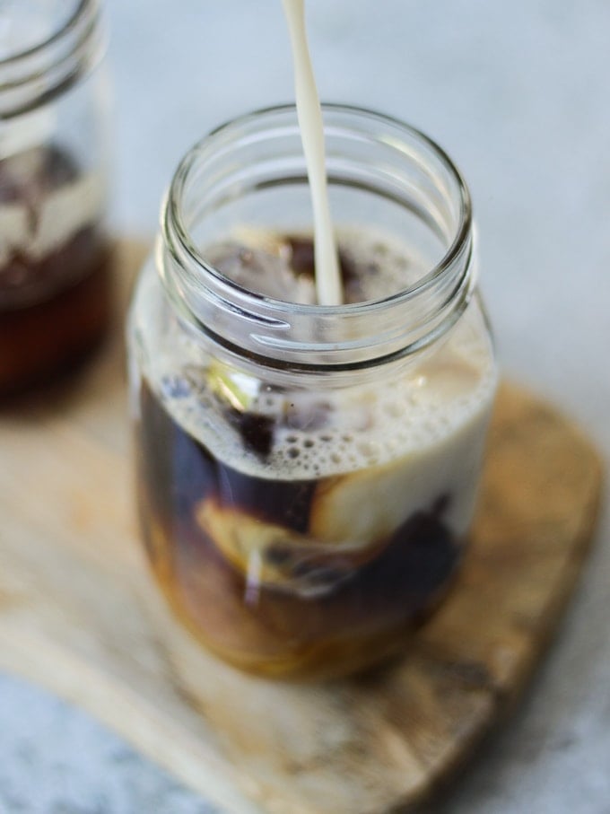 Milk pouring into iced coffee in a glass jar mug on a wooden board.