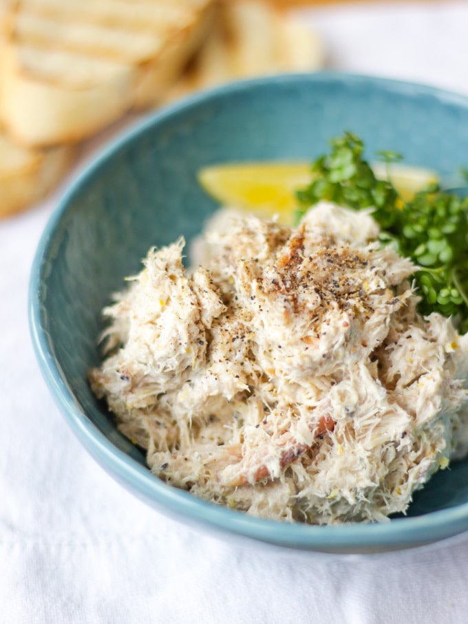 Pile of Smoked Mackerel pate in a green bowl with cress and a wedge of lemon with bread slices in the background on a white napkin. Side view photo.