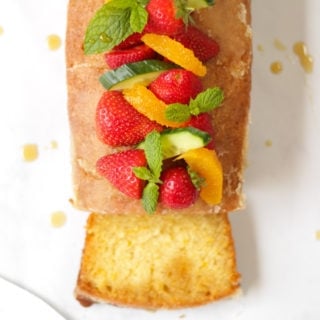 Overhead photo of Orange Drizzle Cake with Pimms. Loaf cake from overhead, topped with strawberries, mint, oranges and cucumber pieces, with slice cut off the cake.