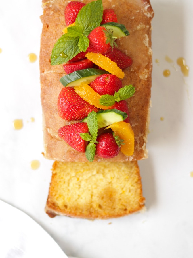 Overhead photo of Orange Drizzle Cake with Pimms. Loaf cake from overhead, topped with strawberries, mint, oranges and cucumber pieces, with slice cut off the cake.