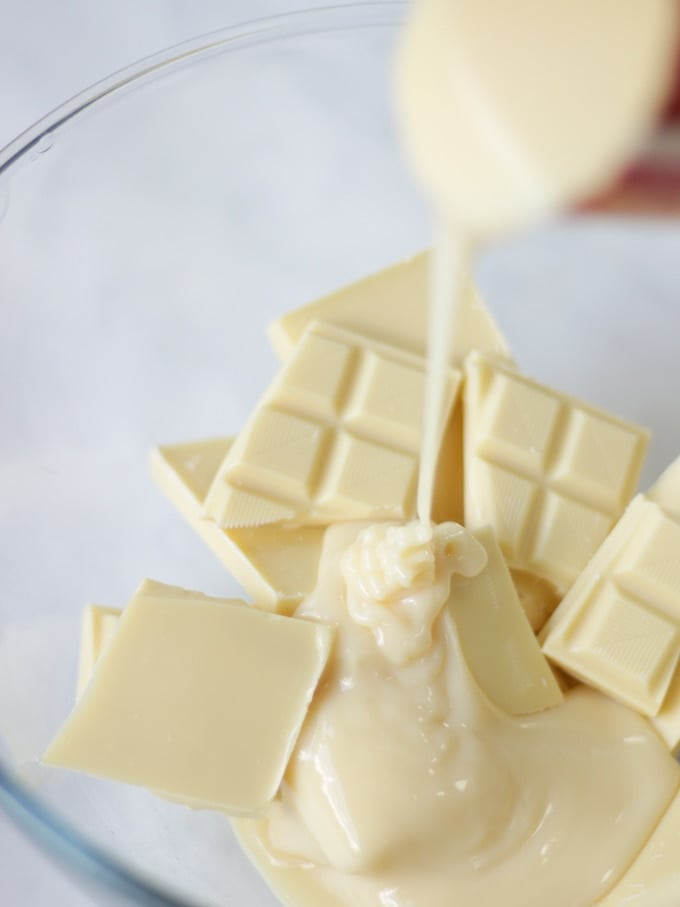 White chocolate pieces in a glass bowl with condensed milk pouring in, in a glass bowl on a white marble background for peanut butter fudge.