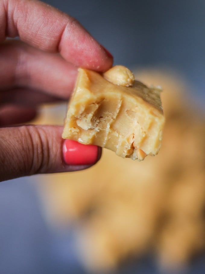 Photo of microwave peanut butter fudge cube in fingers with piece bitten out with salted peanuts on top on grey background.