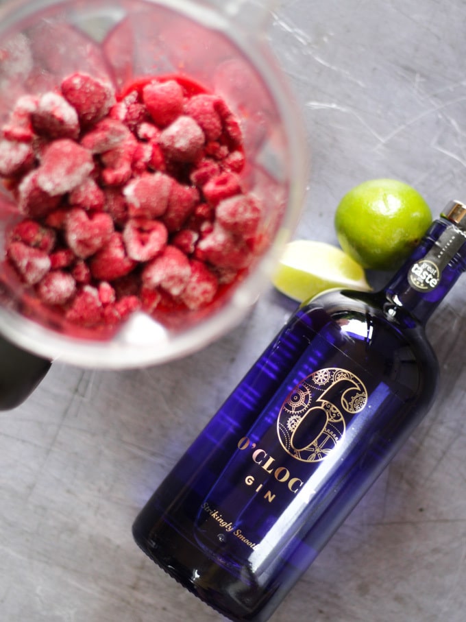 Overhead shot of frozen raspberries in a blender, limes and Waitrose 6 o'clock gin bottle laying down which is blue for Raspberry Gin Frozen Cocktail Recipe.