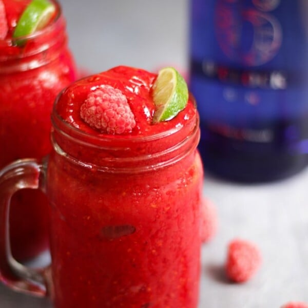 Side photo of raspberry gin frozen cocktails. Adult slushies tipped with raspberries and lime wedges on a grey background.