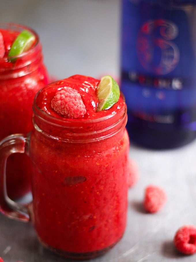 Side photo of raspberry gin frozen cocktails. Adult slushies tipped with raspberries and lime wedges on a grey background.