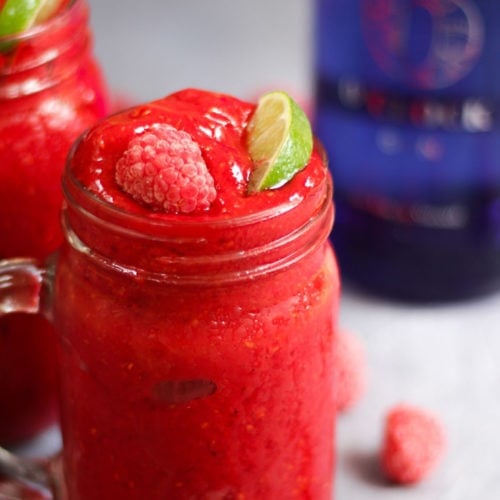 Side photo of raspberry gin frozen cocktails. Adult slushies tipped with raspberries and lime wedges on a grey background.