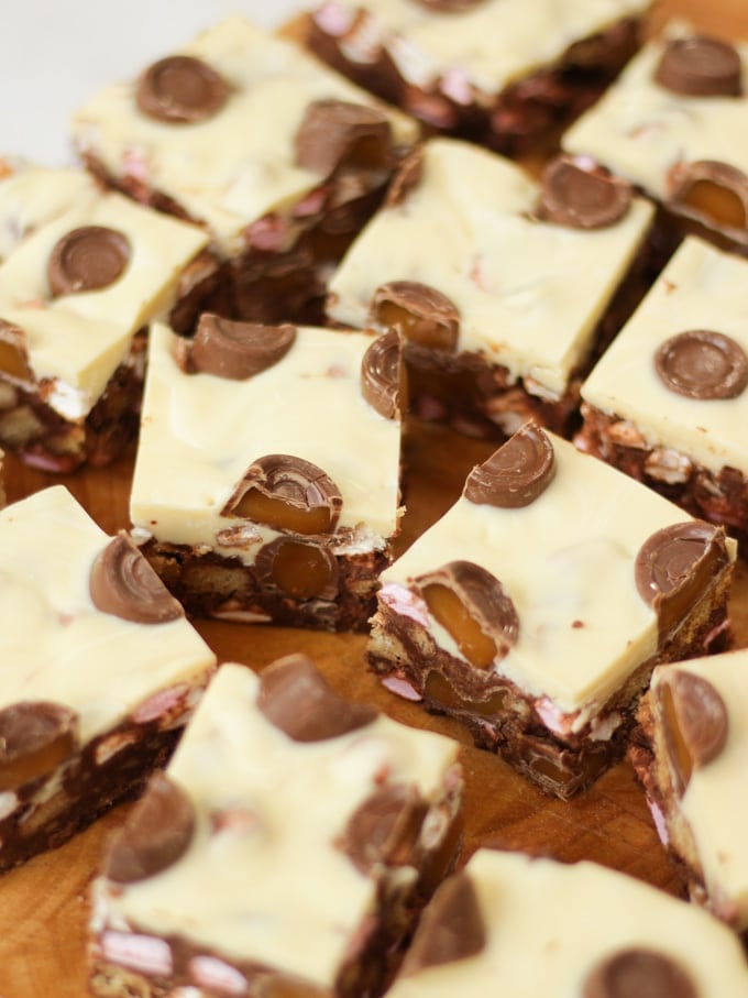 Side angle photo of Rolo Rocky Road Squares made with Roll Rocky Road Recipe, topped with white chocolate and Rolls on a wooden board.