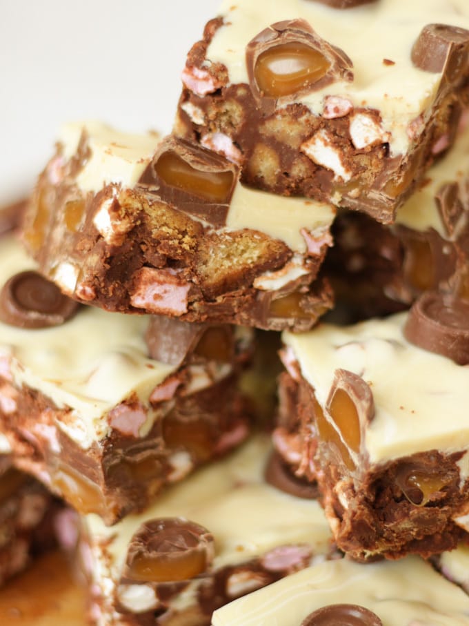 Side angle photo of a pile of Rolo Rocky Road Squares made with Roll Rocky Road Recipe, topped with white chocolate and Rolls on a wooden board.