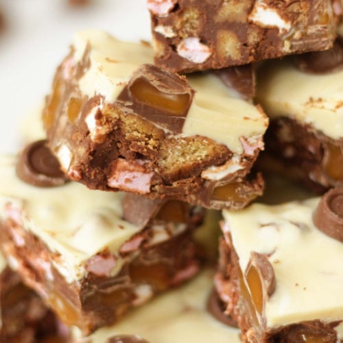 Side angle photo of a pile of Rolo Rocky Road Squares made with Roll Rocky Road Recipe, topped with white chocolate and Rolls on a wooden board.