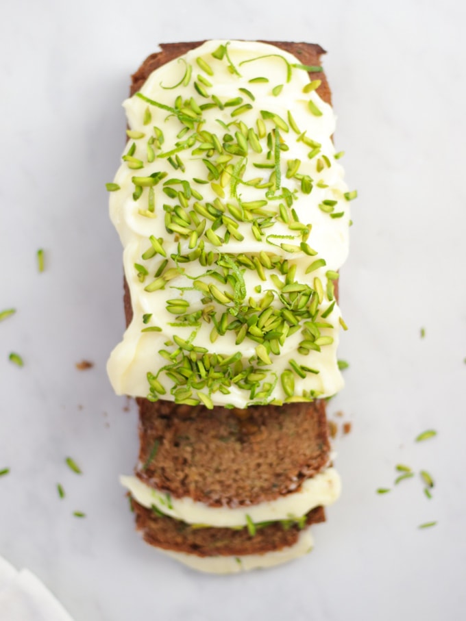 Overhead photo of baked courgette cake loaf topped with cream cheese icing and pistachio nut slices on white marble background.
