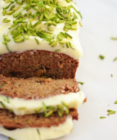 Side angle photo of baked courgette cake loaf topped with cream cheese icing and pistachio nut slices on white marble background.