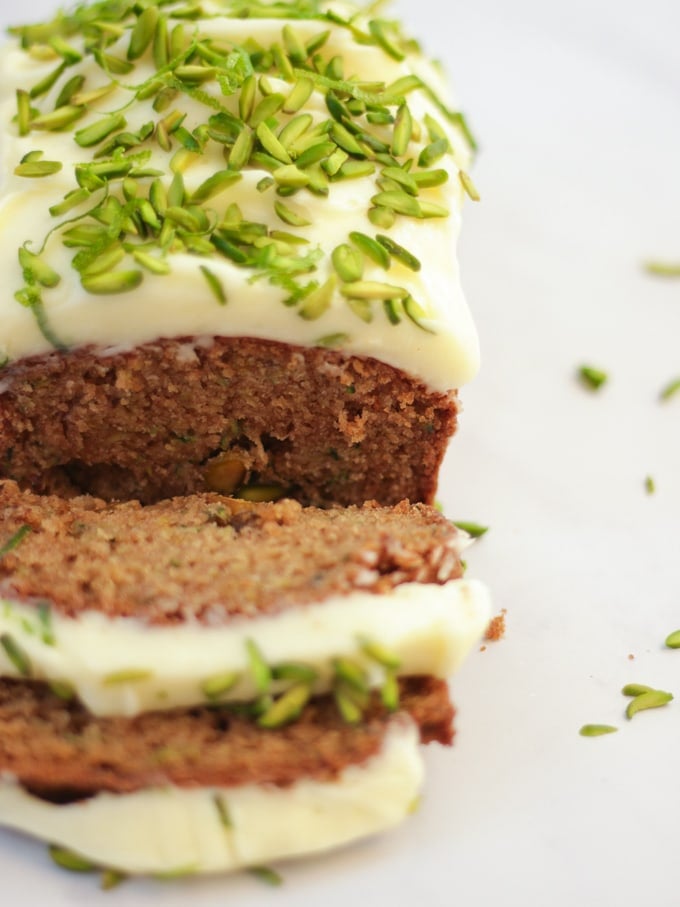 Side angle photo of baked courgette cake loaf topped with cream cheese icing and pistachio nut slices on white marble background.