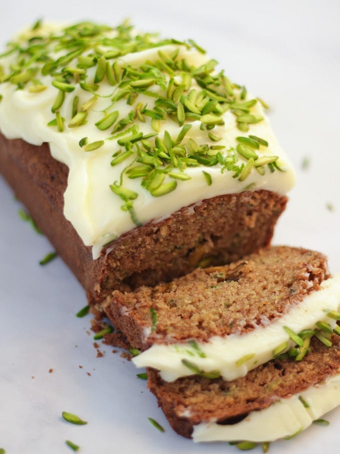 Side angle photo of baked courgette cake loaf topped with cream cheese icing and pistachio nut slices on white marble background.