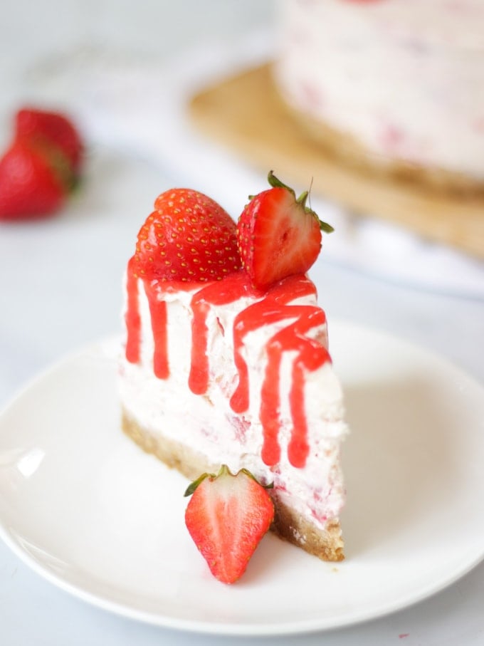 Close up of slice of strawberry cheesecake with Prosecco with strawberry coulis drizzle on a white plate with cheesecake in the background on white marble surface.