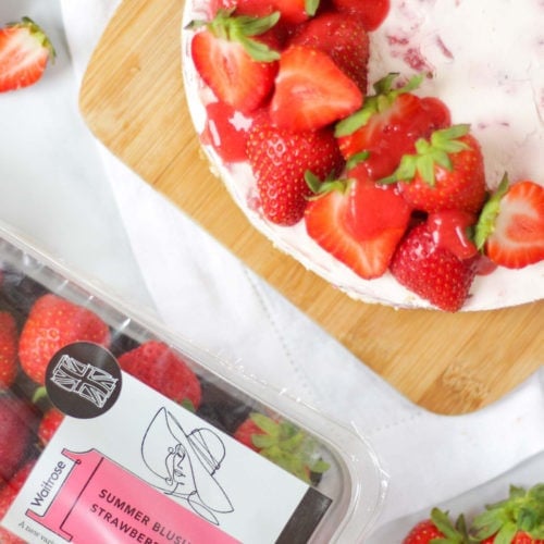 Overhead photo of strawberry cheesecake top with halves strawberries and strawberry coulis drizzle on wooden board and white marble background with pack of Waitrose strawberries ready for recipe in photo too.