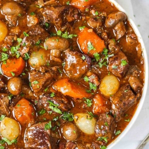 Overhead photo of white bowl of beef bourguignon recipe with beef, carrots, pearl onions and mushrooms showing on white background.