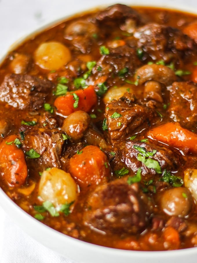 Side photo of white bowl of beef bourguignon recipe with beef, carrots, pearl onions and mushrooms showing on white background.