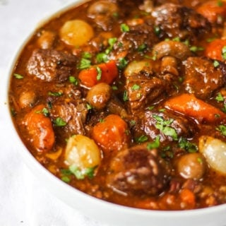 Side photo of white bowl of beef bourguignon recipe with beef, carrots, pearl onions and mushrooms showing on white background.