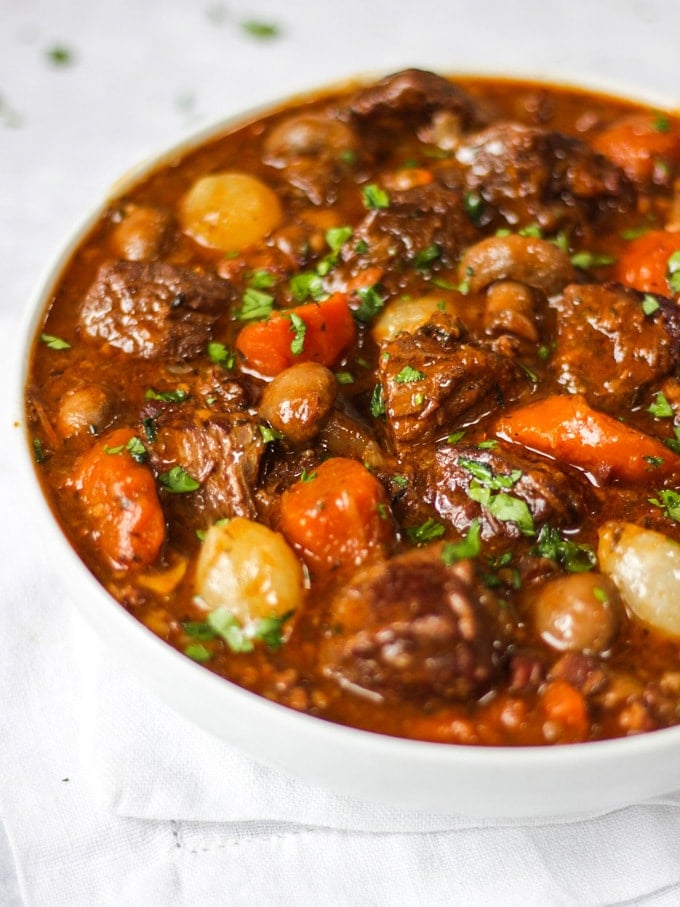 Side photo of white bowl of beef bourguignon recipe with beef, carrots, pearl onions and mushrooms showing on white background.