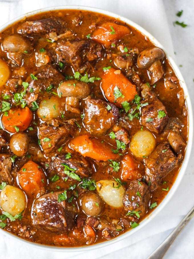 Overhead photo of white bowl of beef bourguignon recipe with beef, carrots, pearl onions and mushrooms showing on white background.