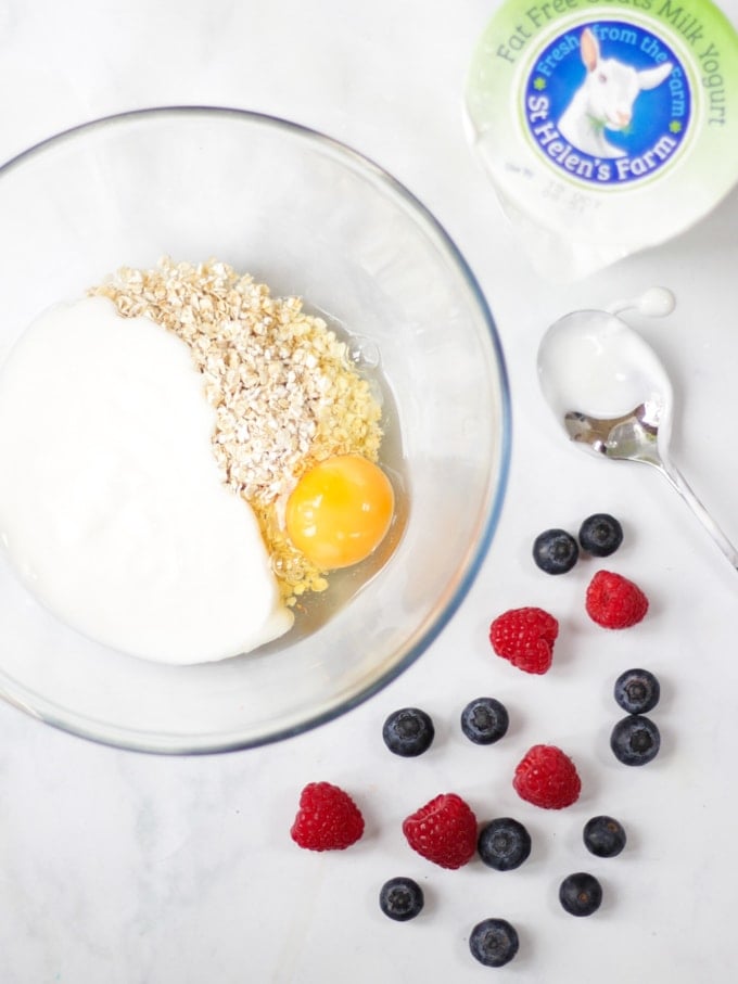 Overhead photo of ingredients for baked oats recipe, yoghurt, oats and eggs in a bowl with raspberries and blue berries on a white background.