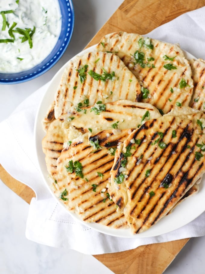 Photo of a plate of flatbreads with griddle lines, on a white plate with white napkin and wooden board, marble background and tzatziki dip in a blog bowl.