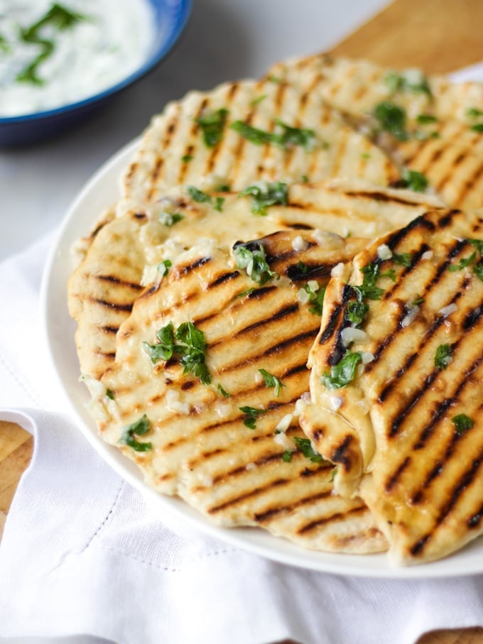 Side photo of a plate of easy flatbreads with griddle lines, on a white plate with white napkin and wooden board, marble background and tzatziki dip in a blog bowl.